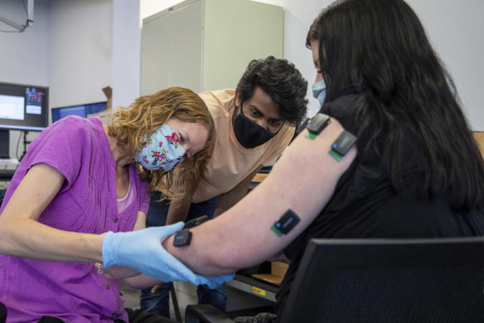 In this photo provided by UPMC and Pitt Health Sciences, occupational therapist Amy Boos, left, and Carnegie Mellon graduate student Nikhil Verma connect sensors on the arm of research participant Heather Rendulic in Pittsburgh on May 19, 2021. A stroke left Rendulic with little use of her left hand and arm, so she volunteered for a first-of-its-kind experiment that stimulates her spinal cord in spots that control upper limb motion. (Tim Betler/UPMC and Pitt Health Sciences via AP)