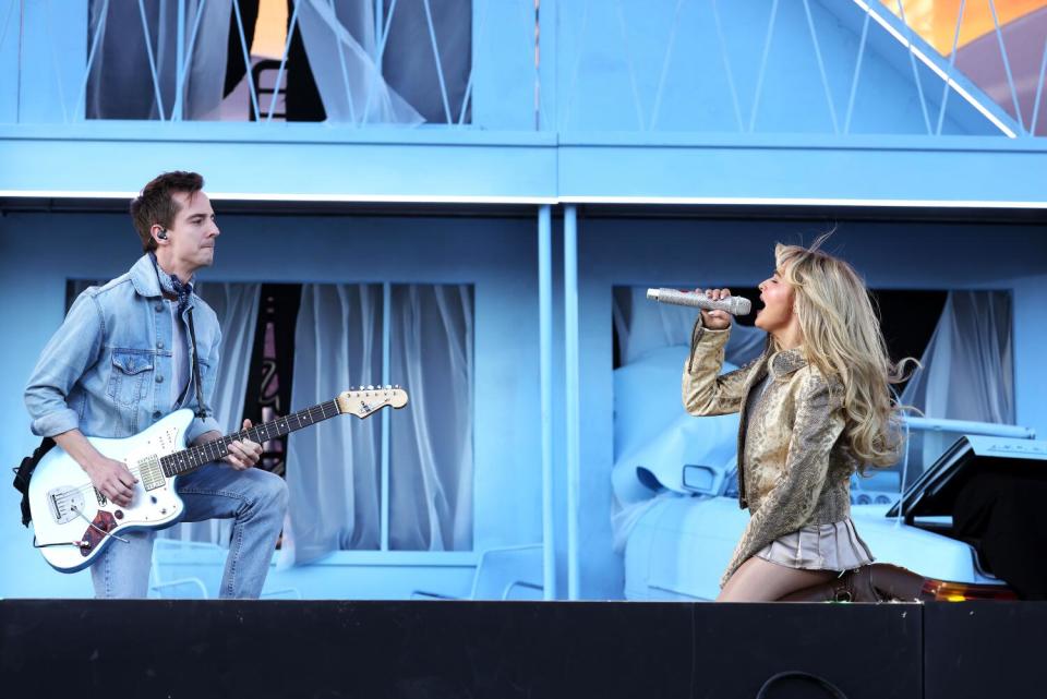 A woman signs into a microphone while another person plays electric guitar in front of a building.