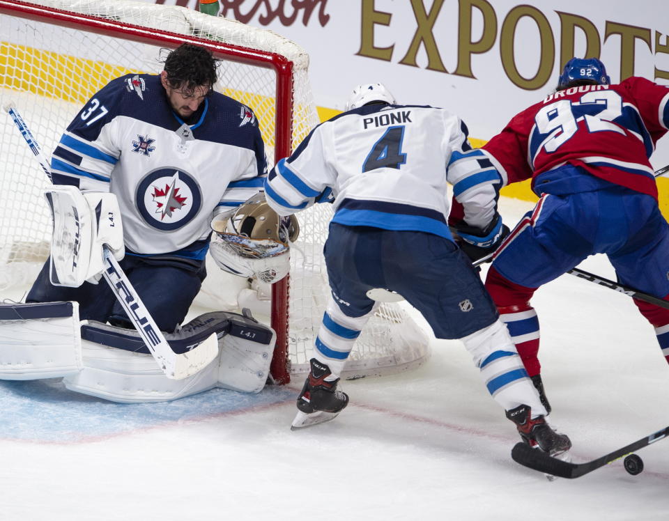 Winnipeg Jets goaltender Connor Hellebuyck (37) catches his mask after making a save as Montreal Canadiens' Jonathan Drouin (92) and Jets' Neal Pionk (4) battle for the rebound during the second period of an NHL hockey game, Thursday, April 8, 2021 in Montreal. (Ryan Remiorz/The Canadian Press via AP)
