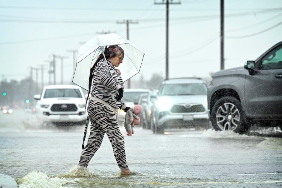 A woman, shielded with an umbrella, carefully crosses the flooded intersection at Foothill Blvd and Cactus Ave in Rialto, Calif., during a downpour, Monday, Feb. 5, 2024. (Watchara Phomicinda/The Orange County Register via AP)