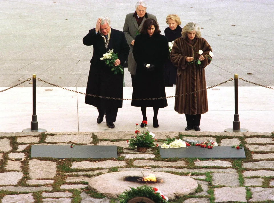 Sen. Ted Kennedy and other members of the Kennedy family, in winter coats, look over a chained-off area with an eternal flame at the grave of his brother John.