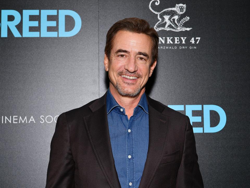 Dermot Mulroney wearing a dark-brown blazer and a blue shirt with a background