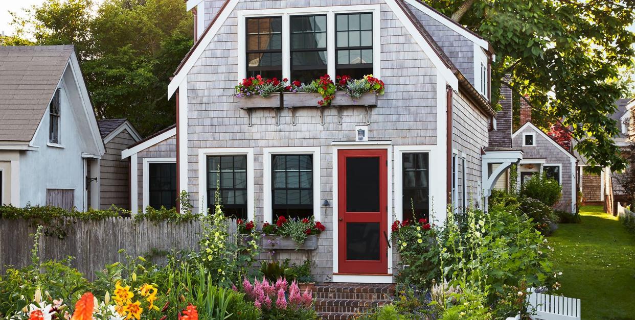 the exterior of a cedar shingled two story cottage with window boxes with red flowers, a red screen door, and brick steps, a white picket fence with roses surrounds tall orange flowers, yellow day lilies, pink foxglove