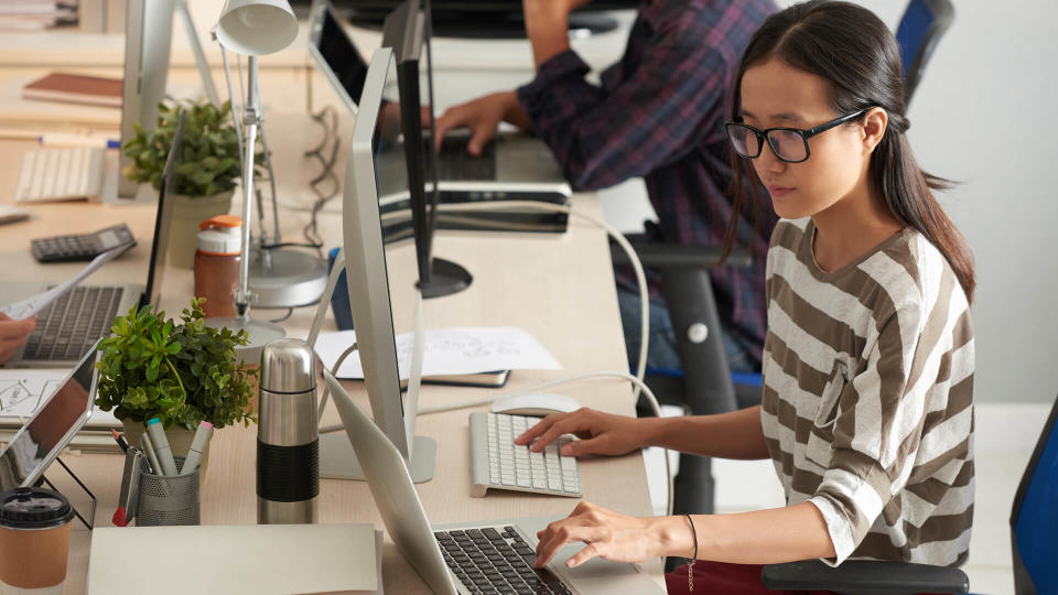 Working process at modern open plan office: pretty young manager in casualwear analyzing statistic data while using modern computer and laptop, profile view.