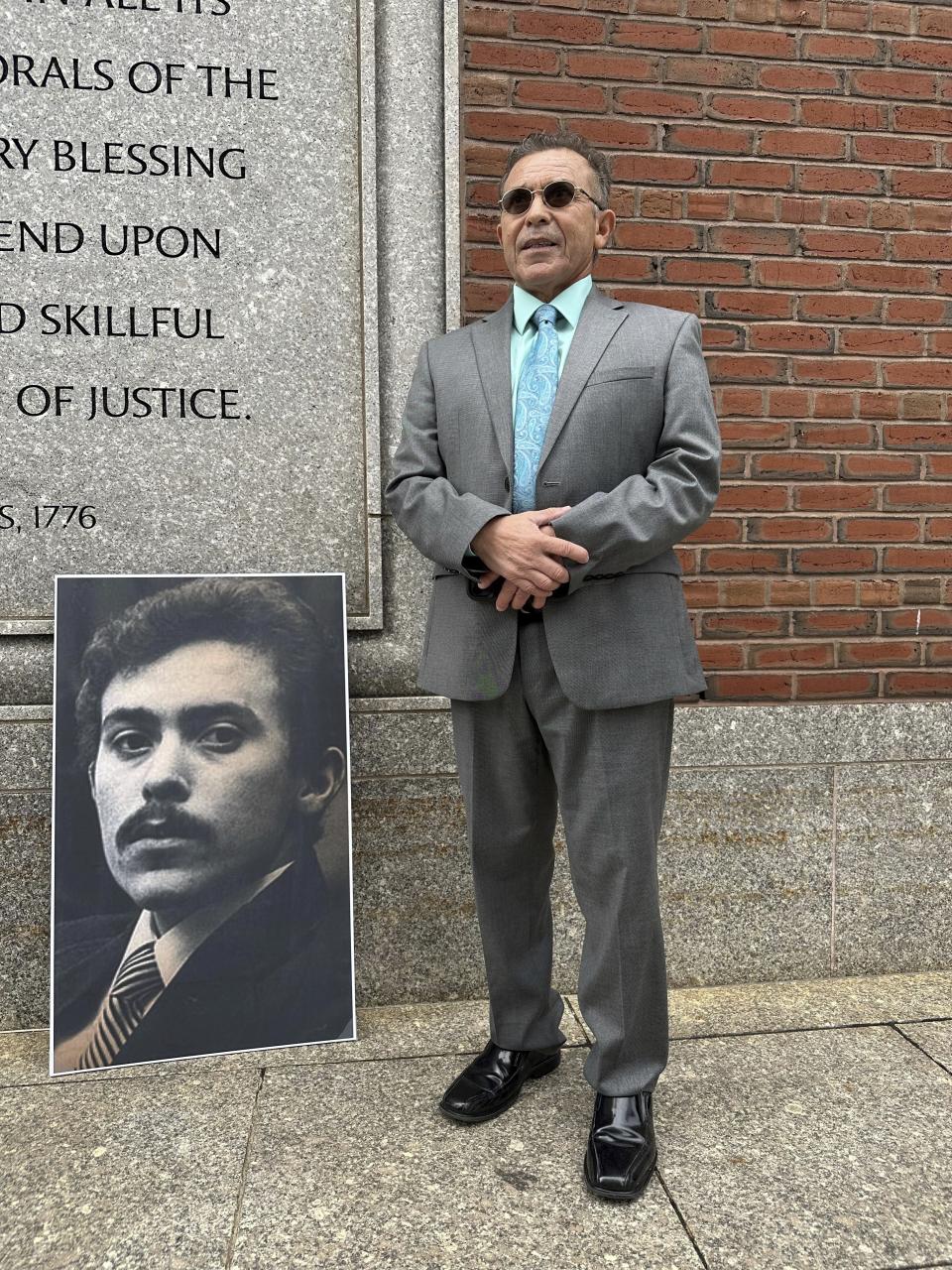 Victor Rosario, 65, stands next to a photo of himself at age 24 when he was on trial in 1982 for arson, Wednesday, May 3, 2023, outside the federal courthouse in Boston. Rosario, who spent 32 years in prison after he was wrongfully convicted of setting a fire that killed eight people, will receive $13 million from the city of Lowell, Mass., in a settlement of a lawsuit he brought against the city in 2019. (AP Photo/Mark Pratt)