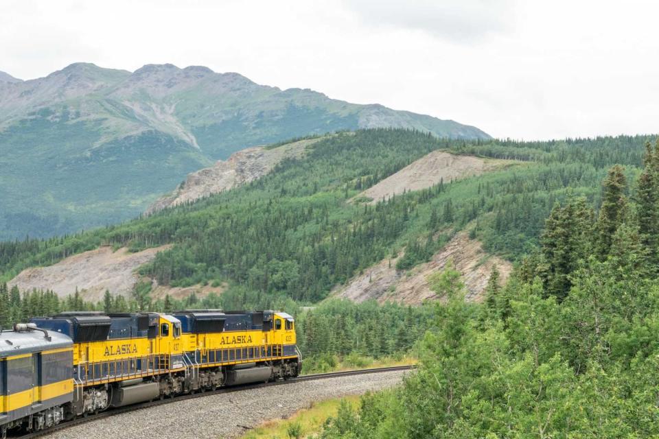 Yellow train with "Alaska" on the side driving through tree-covered mountains