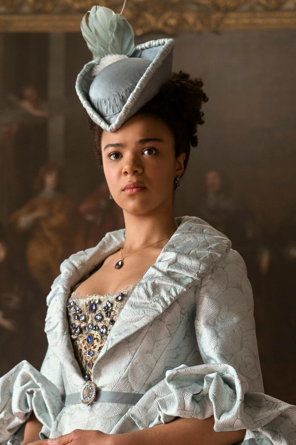 Actress in a period drama wearing an ornate dress with ruffles and a feathered hat, in a room with classic paintings