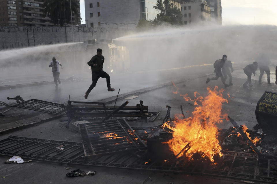 Anti-government protesters run from police spraying water cannons where a street barricade burns, set by demonstrators, in Santiago, Chile, Tuesday, Oct. 29, 2019. Chileans gathered Tuesday for a 12th day of demonstrations that began with youth protests over a subway fare hike and have become a national movement demanding greater socio-economic equality and better public services in a country long seen as an economic success story. (AP Photo/Rodrigo Abd)