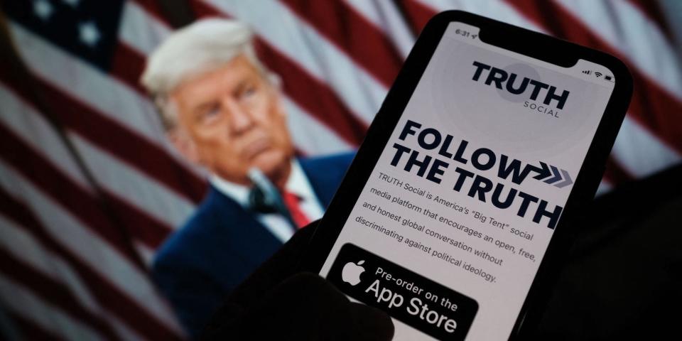 A photo illustration showing a person checking the app store on a smartphone for "Truth Social", with a photo of former President Donald Trump in the background.