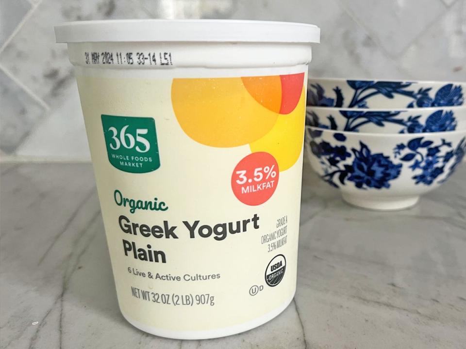 A light-yellow container of 365 organic plain Greek yogurt with a red, orange, and yellow design on the packaging. The container sits on a gray marble countertop with blue-and-white bowls in the backgrounds