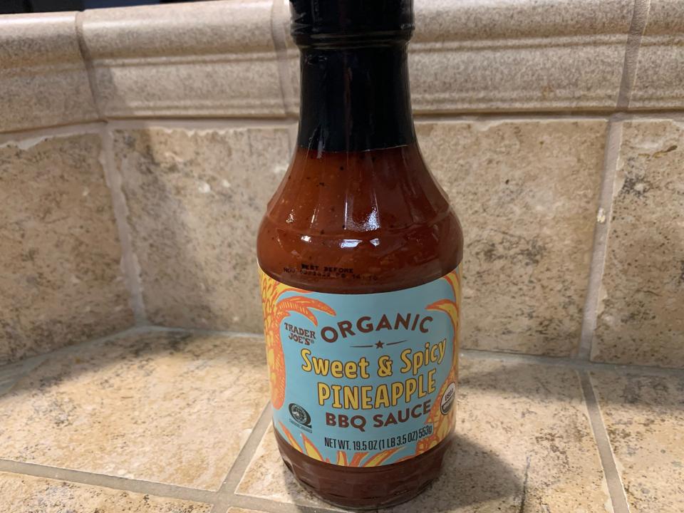 Trader joe's pineapple barbecue sauce in the original clear, blue, and orange bottle against beige tile