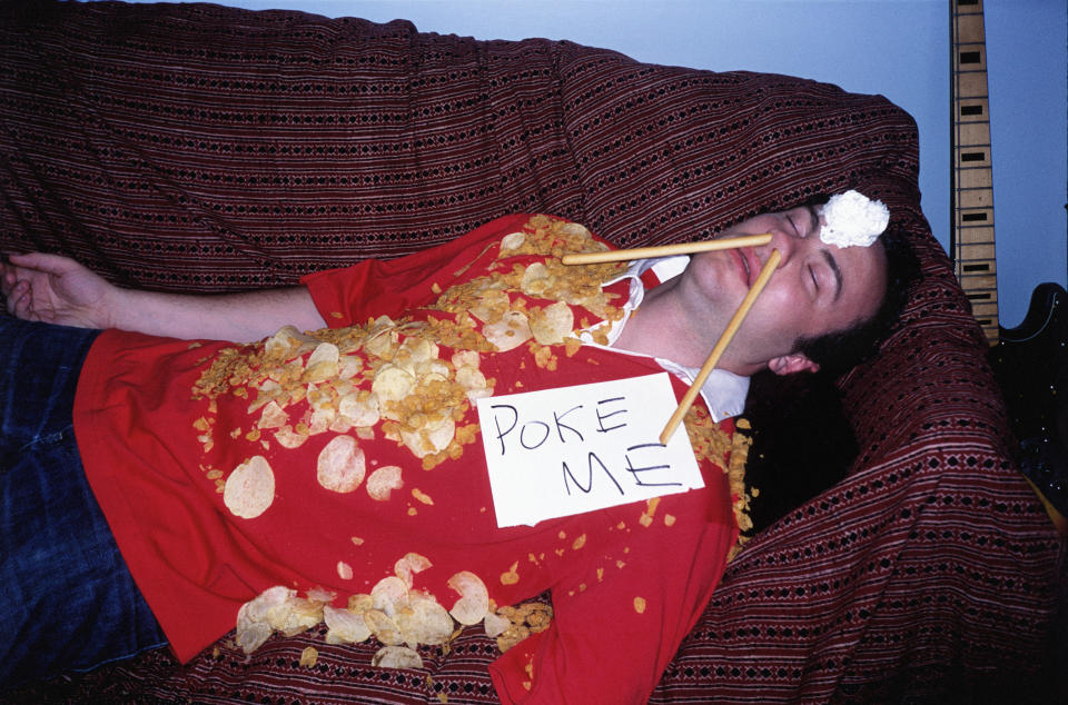 A very drunk person lying on a couch with potato chips all over their T-shirt, breadsticks emerging form their nostrils, and a "Poke Me" sign on their chest