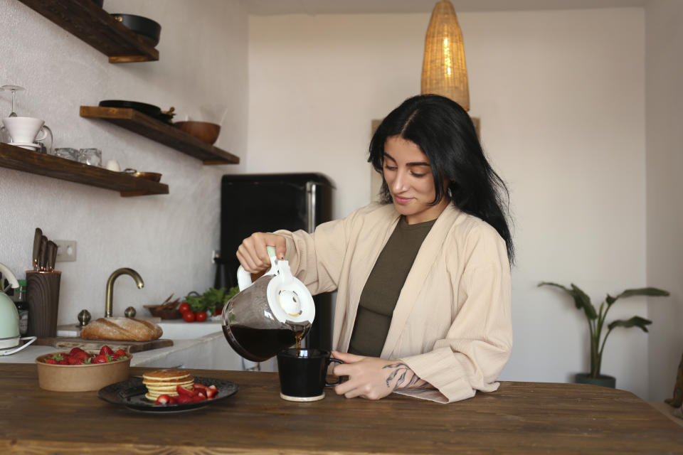 A woman in a kitchen pours coffee from a coffeepot into a mug.