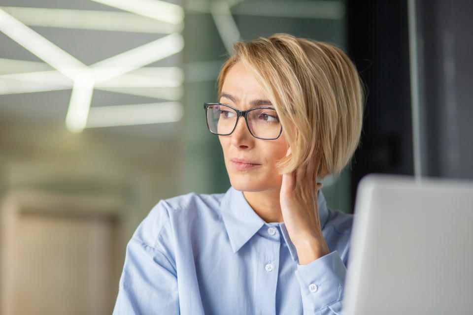 Woman in glasses looking thoughtfully at a laptop screen in an office setting