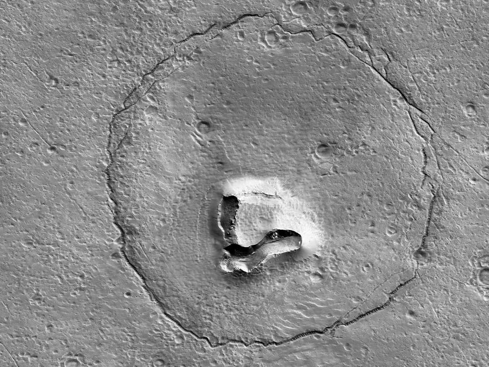 A black and white picture taken by Mars Reconnaissance Orbiter of a peculiar rock formation. A crack runs in a circle around a hill and two craters, making it look like the face of a bear.