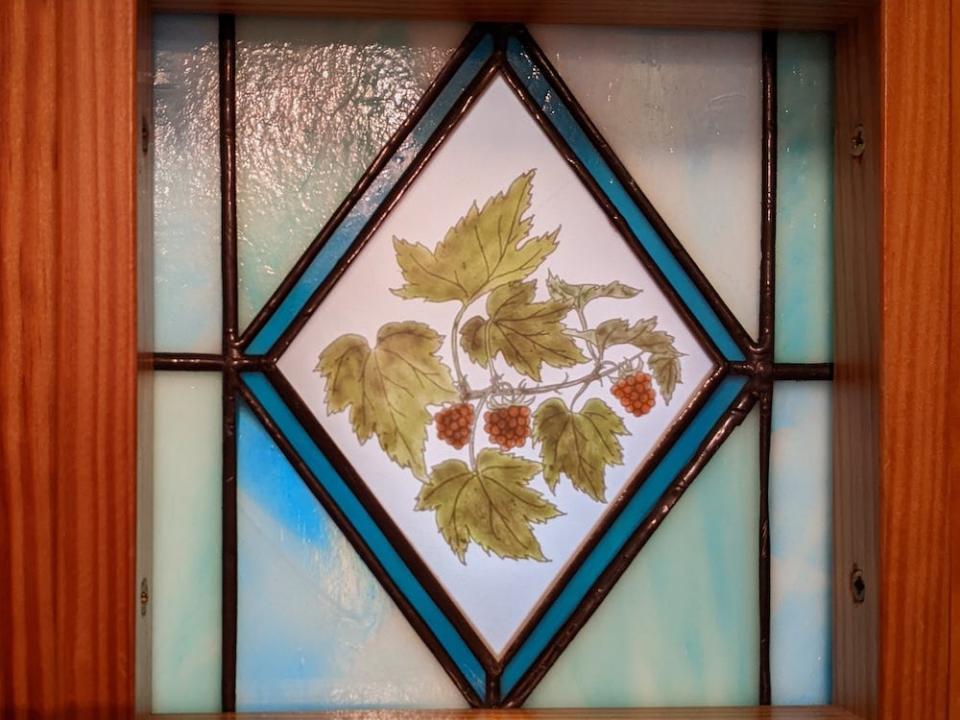 A wooden door with a stained glass window. The glass is light blue with a blue diamond border and an image of raspberries on a vine with leaves.