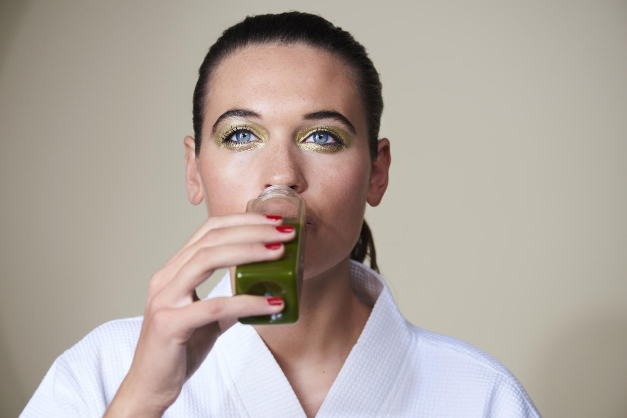 A model with clear skin, her hair tied back and wearing a white robe, drinks a bottle of green juice