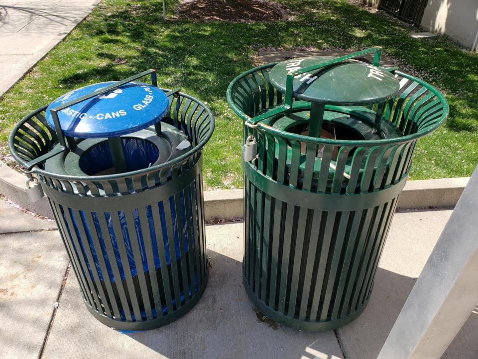 Two plastic trash cans with covers on a sidewalk next to a patch of grass.