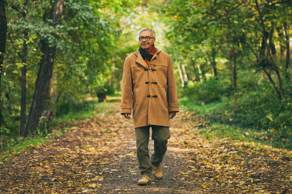 Older man in a coat, walking on a paved path surrounded by early-autumn woods