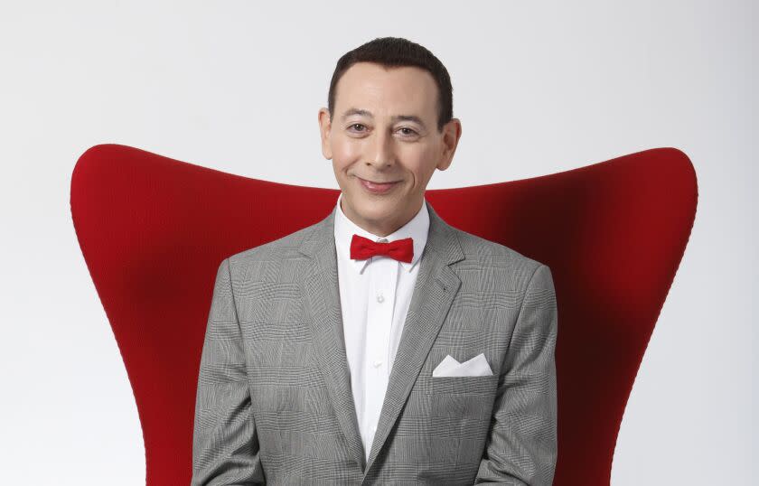 Paul Reubens sitting in a red chair and smiling in a gray suit and red bowtie