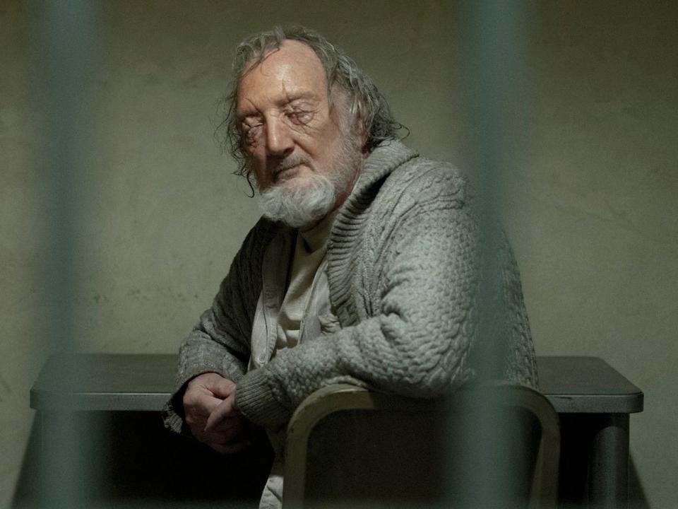 An older man with scarred eyes looks out between his prison cell bars.