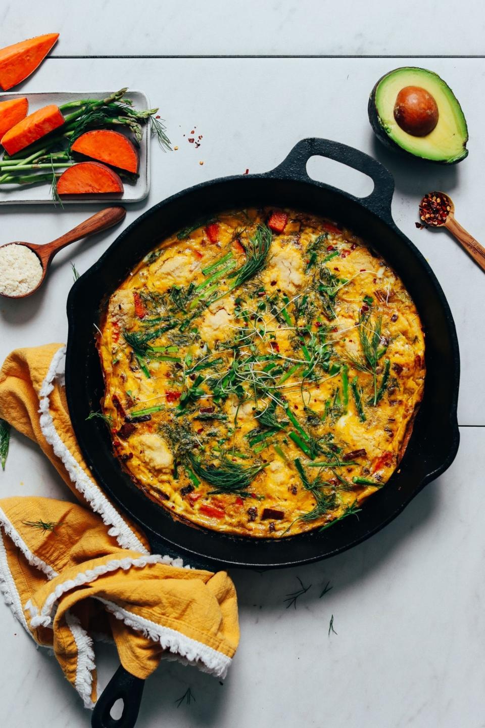 A skillet with a cooked frittata garnished with herbs, surrounded by raw ingredients