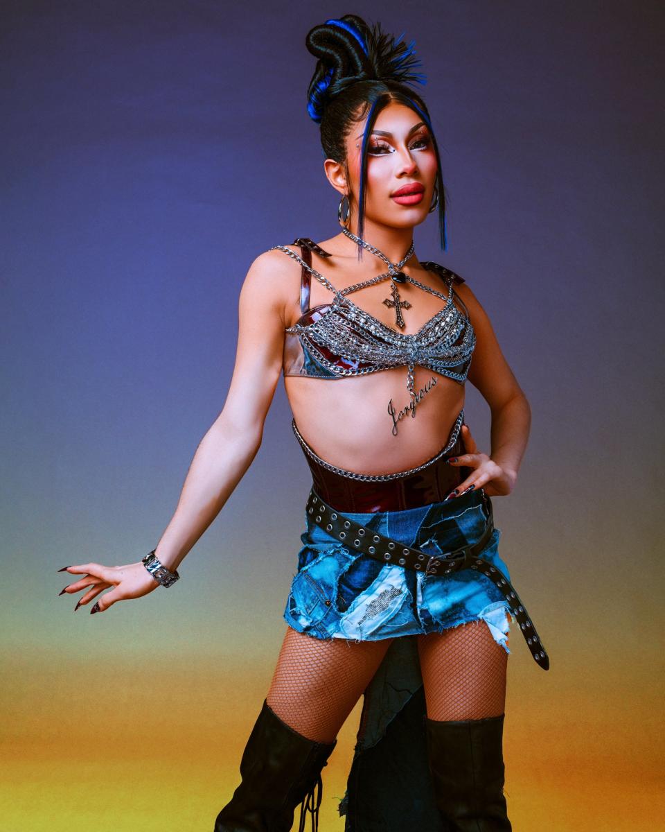 Jorgeous poses in an elaborate outfit featuring a bejeweled bra top, denim mini skirt with chain details, fishnet stockings, and thigh-high black boots