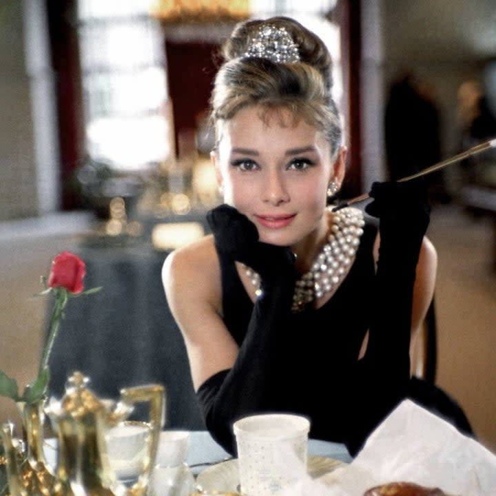 Audrey at table with coffee and pastry, wearing a dress, gloves, and pearl necklace