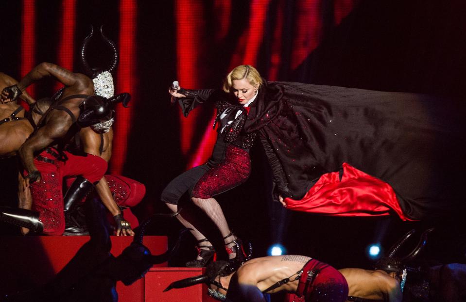 Madonna in a red short jumpsuit and a long black and red cape. The cape is flying in the air and she's falling backwards on stairs.