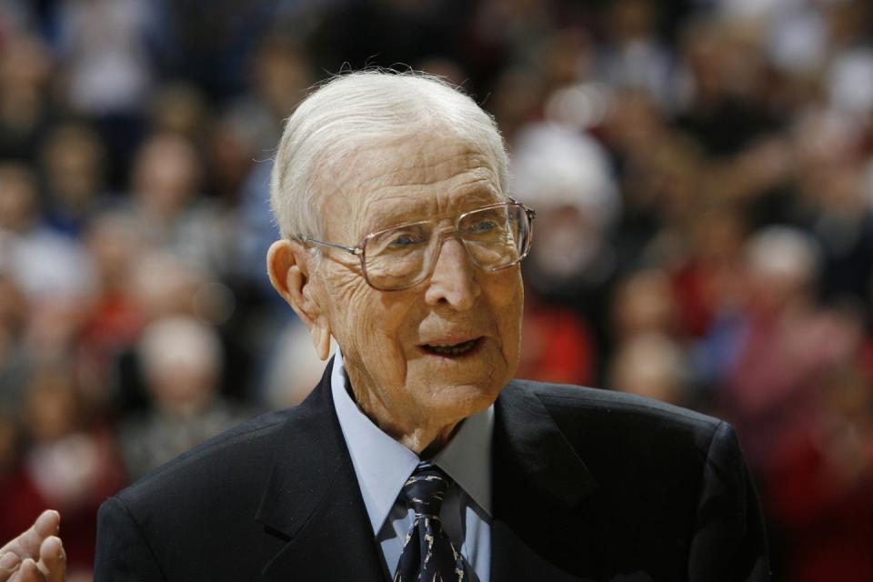 john wooden wearing a black suit and blue shirt, and glasses, smiling and looking off camera at a basketball game