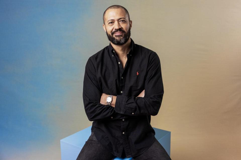 Cord Jefferson sits in a dark outfit with his arms crossed while sitting on a cube and smiling.