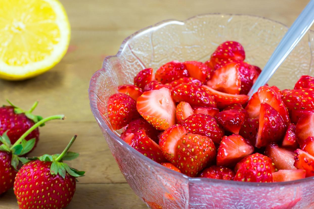 Simple macerated strawberries in a glass fruit bowl with a spoon on a wooden table with strawberries and a lemon