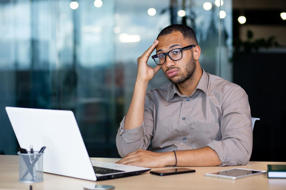 Man in office looks stressed with hand on forehead, sitting at desk with laptop and smartphone