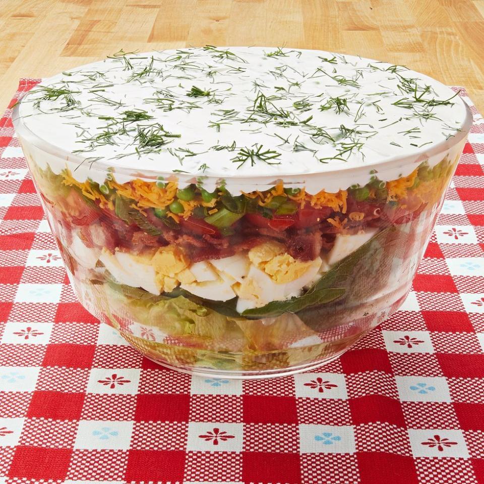 layered salad in big glass bowl topped with white dressing on red checkered linen