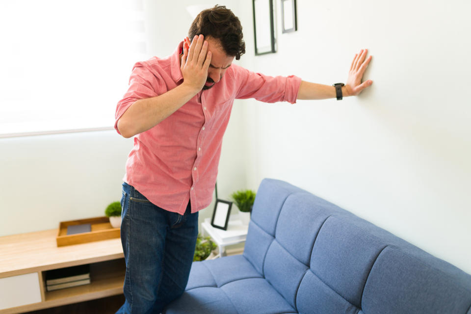 Man leaning against a wall in a living room, touching his head to relieve his dizziness.