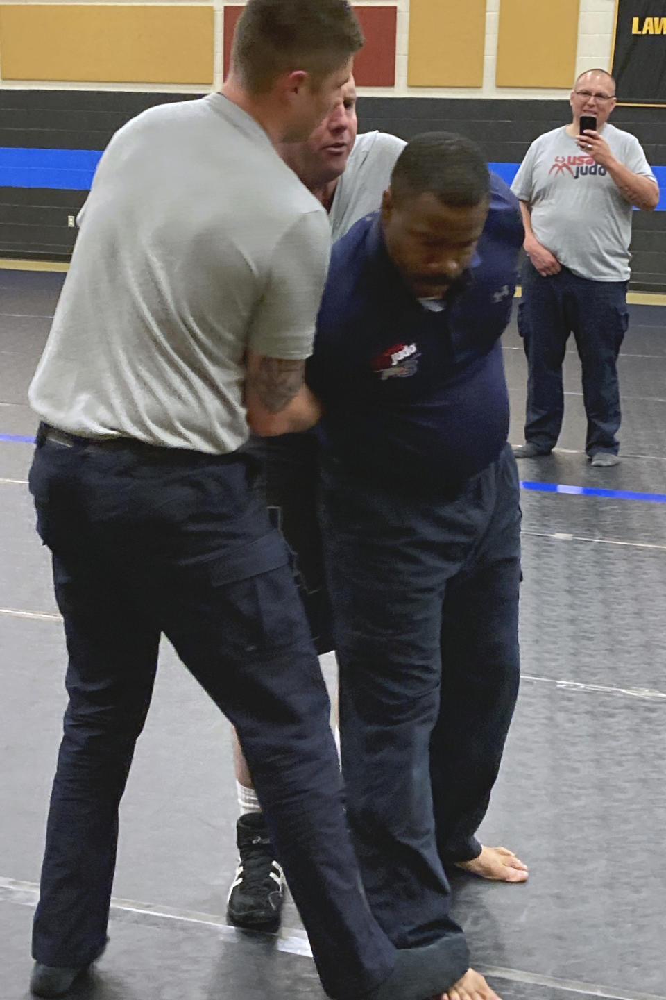 Matthew Maestas, left, of the Torrington, Wyoming Police Department and Ryan Rhoades, obscured at center, of the Meridian, Idaho Police Department practice a judo hold on instructor Taybren Lee of the LA Police Department during a training workshop on March 9, 2021, in Douglas, Wyoming. John Richard of the Glen Rock Wyoming Police Department captures the action with his camera phone. USA Judo is sponsoring training sessions for police departments throughout the country in hopes of preventing the use of deadly force when officers encounter possible suspects on the street. (AP Photo/Eddie Pells)