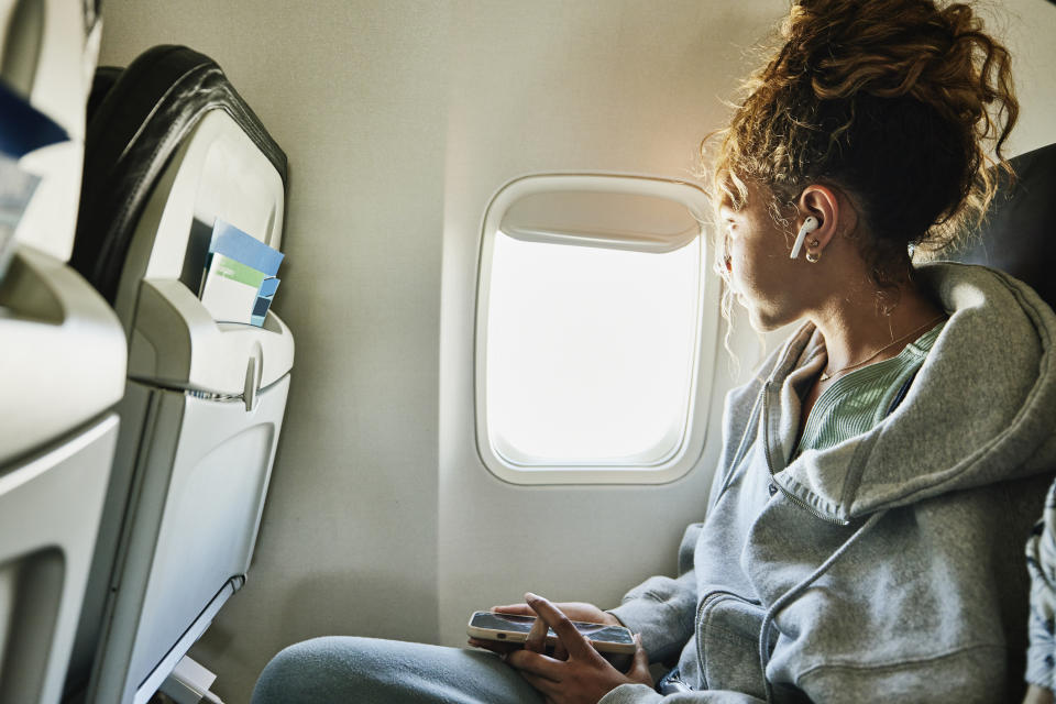 Woman with curly hair wearing earbuds, sitting by an airplane window, looking outside, holding a smartphone in her lap, dressed in casual loungewear