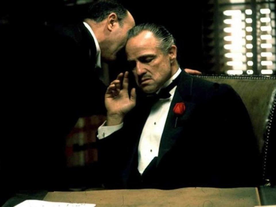 A man whispering into Brando's ear in a dark office with window shutters letting in a little bit of light.