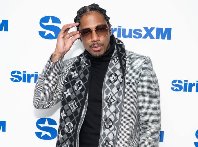 Nick Cannon poses in a checkered suit with a patterned scarf at a SiriusXM event backdrop, adjusting his sunglasses