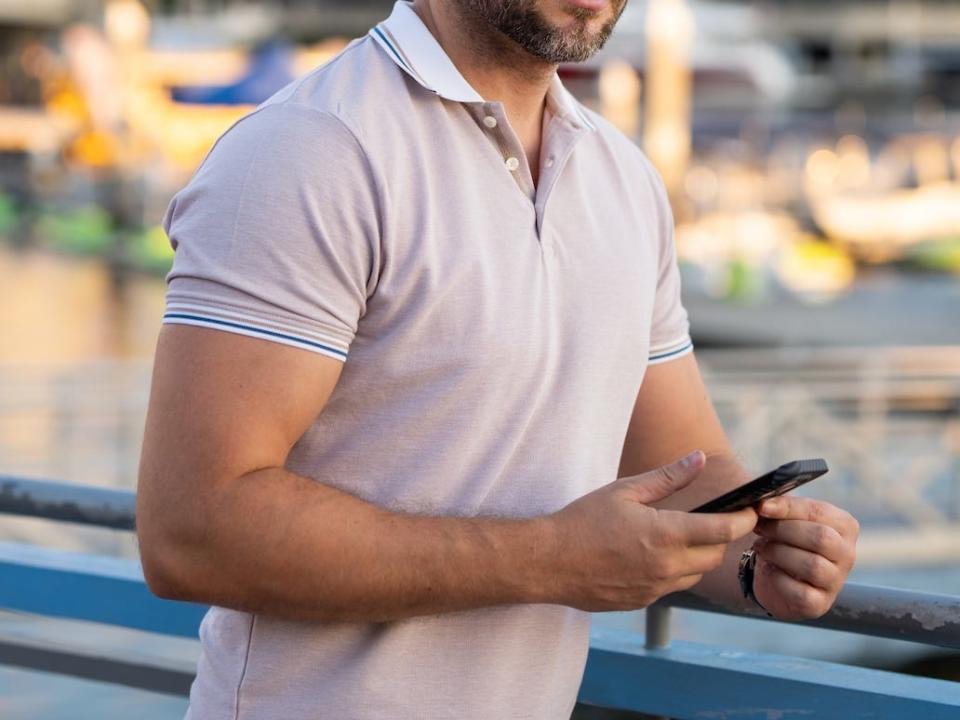 man wearing a pink polo shirt checking his phone on the sidewalk by a pier