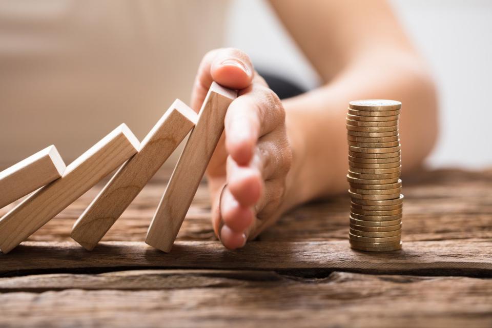 A hand stopping falling dominos from overturning a stock of coins.