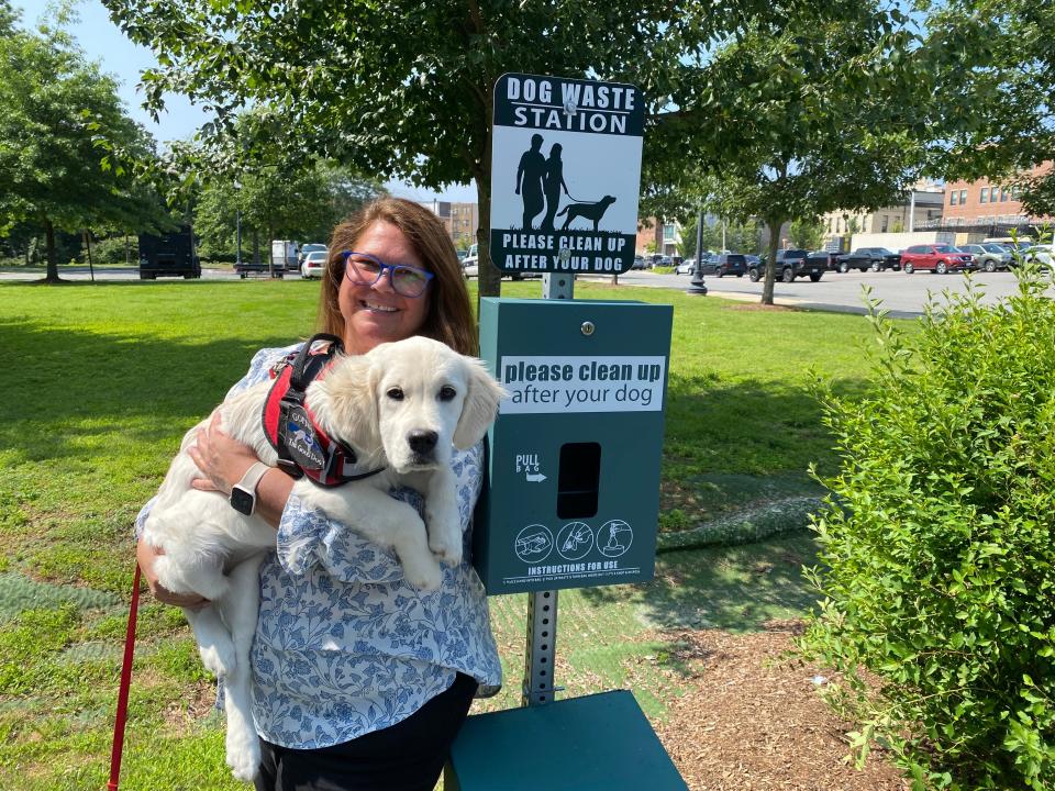 Taunton PD's comfort dog, Maggie, being held by her handler, Katrina Lee, Co-Response Clinician for the Police Department. They are standing next to one of 4 dog waste stations installed around the downtown area. This dog waste station is located in the Mill River park/walking path area, which is adjacent to Spring St. and the parking lot behind the police station. Photo taken July 26, 2023.