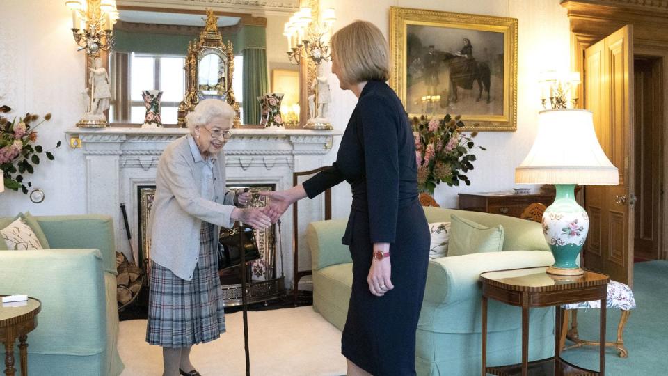 queen elizabeth ii shakes hands with liz truss as both women stand in a living room, elizabeth wears a gray cardigan, blue shirt, and plaid skirt, truss wears an all black skirt suit, the room has green carpet, two green couches and a fireplace with several decorations