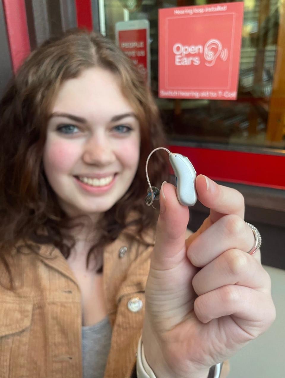 Isabella Froh holds a hearing aid in front of Johnston's Bakery in Sheboygan, Wis., where she helped get an Open Ears hearing loop installed.