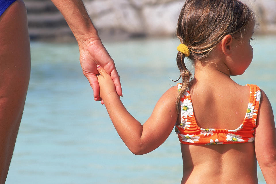 A child in a floral swimsuit holds an adult's hand near water. Names of persons in the image are unknown