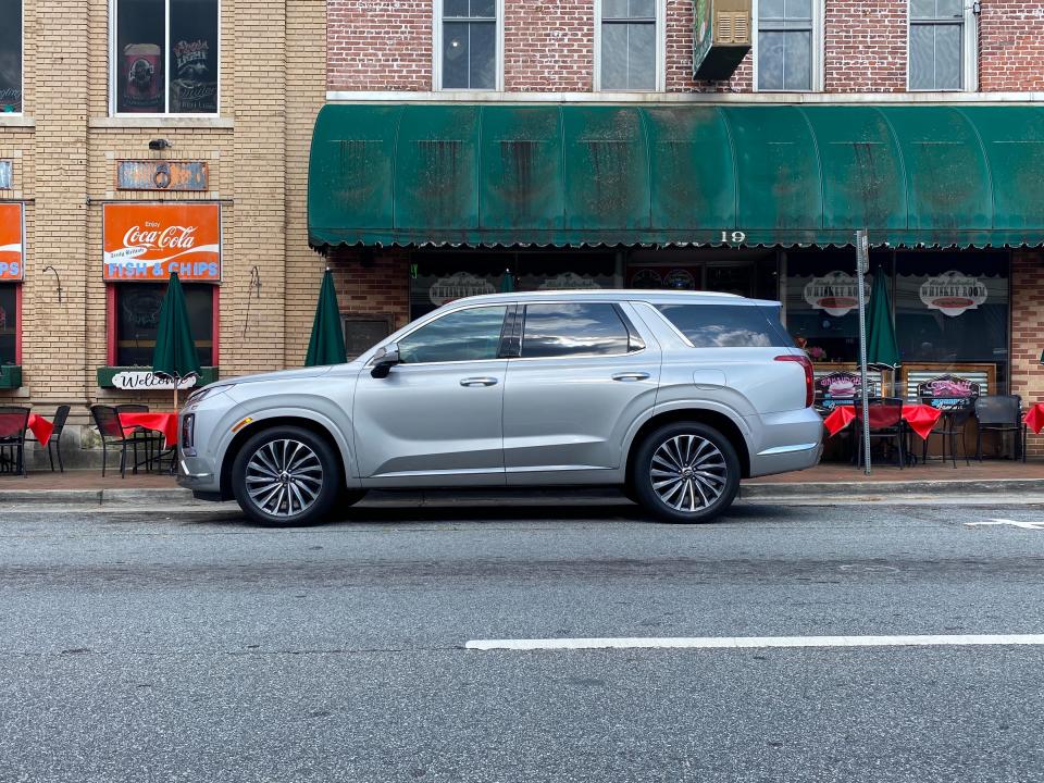 The side view of a silver Hyundai Palisade SUV parked on the street in front of a restaurant.