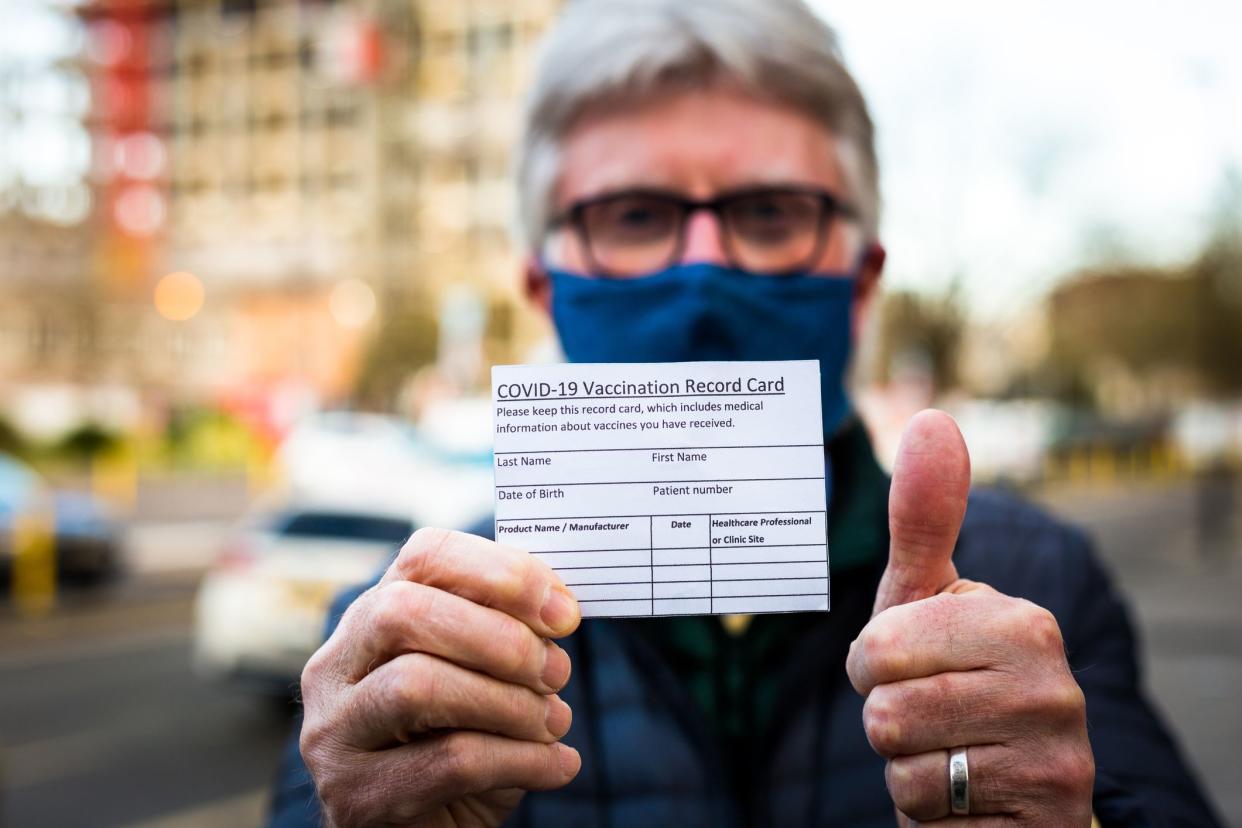 Close up color image depicting a senior man in his 70s holding a card outdoors in the city with details of his covid-19 vaccination. Focus on the card in the foreground while the man is defocused beyond. Room for copy space.