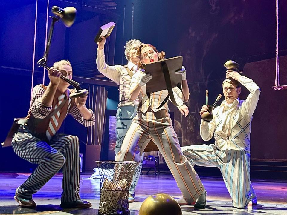 Cirque du Soleil performers on a stage, dressed in pin-striped costumes with heavy white makeup and holding various props. In front of them is a wastebasket with paper in it. They are lit by a purple light.