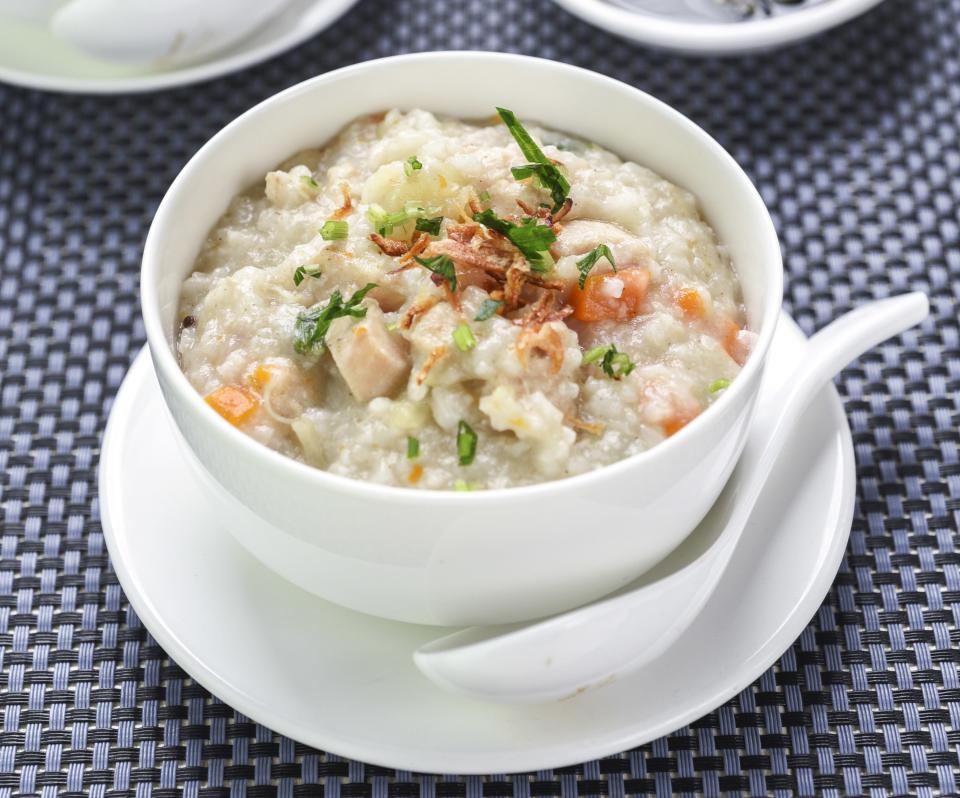 Indonesian congee with shredded chicken is in a white bowl on a white plate. A white spoon is on its side on the white plate and touching the white bowl. A mat of two different blue hues is underneath the plate