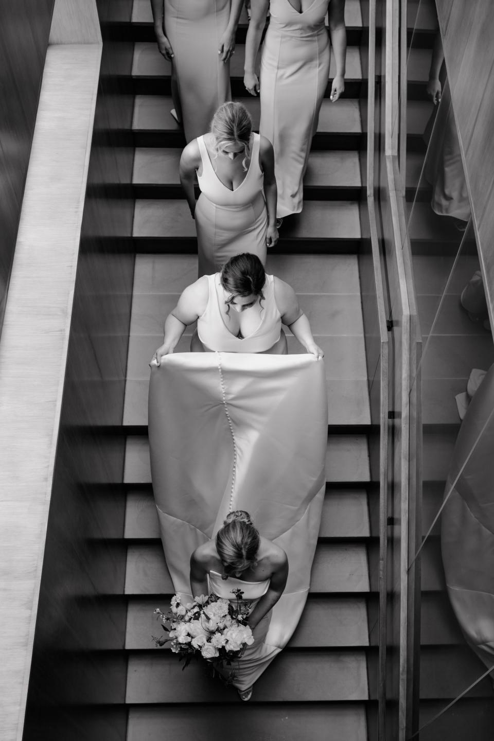 A bride walks down a staircase as her bridesmaids carry her dress and follow her.
